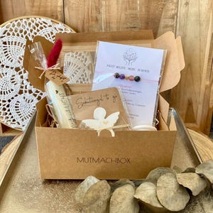 Könnte beinhalten: Eine Geschenkbox mit dem Text "MUTMACHBOX" enthält ein Armband, eine Kerze und andere kleine Gegenstände. Die Box steht auf einem silbernen Tablett und einer Holzscheibe, mit einem Holztablett im Hintergrund. Eine weiße Spitze und getrocknete Blätter sind ebenfalls sichtbar.
