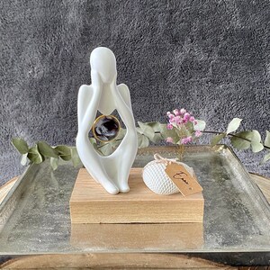 K&ouml;nnte beinhalten: Wei&szlig;e Keramikskulptur einer Person, die sich das Gesicht h&auml;lt, mit einer runden, durchsichtigen Glaskugel im Inneren der Skulptur. Die Skulptur steht auf einem Holzsockel und einem silbernen Tablett, das auf einer Holzscheibe liegt.