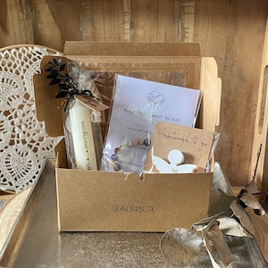 Könnte beinhalten: Eine braune Pappschachtel mit der Aufschrift TRAUERBOX, die eine weiße Kerze mit Text, ein kleines Perlenarmband und eine weiße Engelsfigur in einer Kunststoffhülle enthält. Die Box steht auf einem silbernen Tablett.
