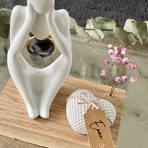 K&ouml;nnte beinhalten: Wei&szlig;e Keramikfigur einer Person, die mit dem Kopf in den H&auml;nden sitzt, auf einem Holzsockel mit einer wei&szlig;en herzf&ouml;rmigen Schachtel mit einem braunen Etikett, auf dem "Emma" steht.