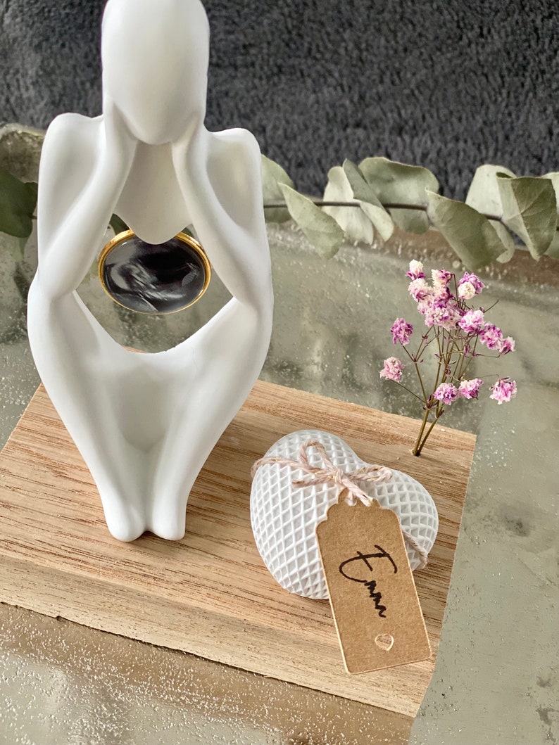 K&ouml;nnte beinhalten: Wei&szlig;e Keramikfigur einer Person, die mit dem Kopf in den H&auml;nden sitzt, auf einem Holzsockel mit einer wei&szlig;en herzf&ouml;rmigen Schachtel mit einem braunen Etikett, auf dem "Emma" steht.