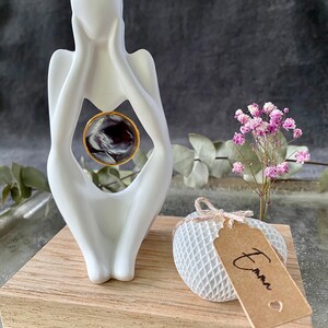 K&ouml;nnte beinhalten: Wei&szlig;e Keramikskulptur einer Person, die sich das Gesicht h&auml;lt, mit einem kleinen runden schwarz-wei&szlig;en Marmor im Inneren der Skulptur. Die Skulptur steht auf einem Holzsockel mit einem wei&szlig;en herzf&ouml;rmigen Objekt mit einem braunen Etikett, auf dem "Emma" steht.