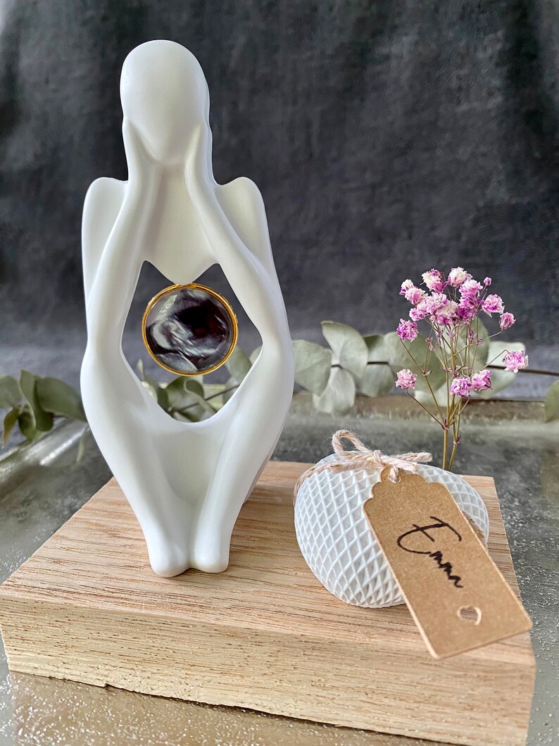 K&ouml;nnte beinhalten: Wei&szlig;e Keramikskulptur einer Person, die sich das Gesicht h&auml;lt, mit einem kleinen runden schwarz-wei&szlig;en Marmor im Inneren der Skulptur. Die Skulptur steht auf einem Holzsockel mit einem wei&szlig;en herzf&ouml;rmigen Objekt mit einem braunen Etikett, auf dem "Emma" steht.