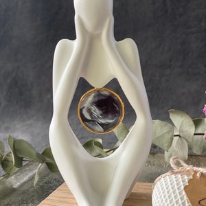 K&ouml;nnte beinhalten: Wei&szlig;e Keramikfigur einer Person mit ineinander verschr&auml;nkten H&auml;nden um eine kreisf&ouml;rmige &Ouml;ffnung. In der &Ouml;ffnung befindet sich ein Schwarzwei&szlig;-Ultraschallbild, das mit einem goldenen Ring umrahmt ist.