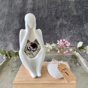 K&ouml;nnte beinhalten: Wei&szlig;e Keramikskulptur einer Person, die ein rundes, schwarz-wei&szlig;es Foto in einem goldenen Rahmen h&auml;lt. Die Skulptur steht auf einem Holzsockel mit einer kleinen wei&szlig;en, netzbedeckten Kugel und einem braunen Etikett mit der Aufschrift "Emma".