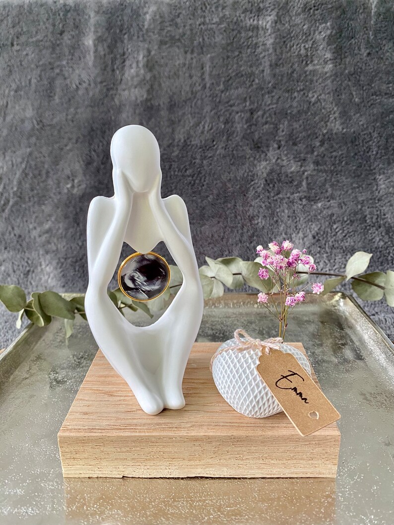K&ouml;nnte beinhalten: Wei&szlig;e Keramikskulptur einer Person, die ein rundes, schwarz-wei&szlig;es Foto in einem goldenen Rahmen h&auml;lt. Die Skulptur steht auf einem Holzsockel mit einer kleinen wei&szlig;en, netzbedeckten Kugel und einem braunen Etikett mit der Aufschrift "Emma".