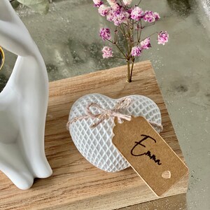 K&ouml;nnte beinhalten: Wei&szlig;e Keramikh&auml;nde halten ein kleines wei&szlig;es Herz mit einem braunen Etikett, auf dem "Emma" steht. Das Herz befindet sich auf einem Holzsockel mit einem kleinen Strau&szlig; rosa und wei&szlig;er Blumen.