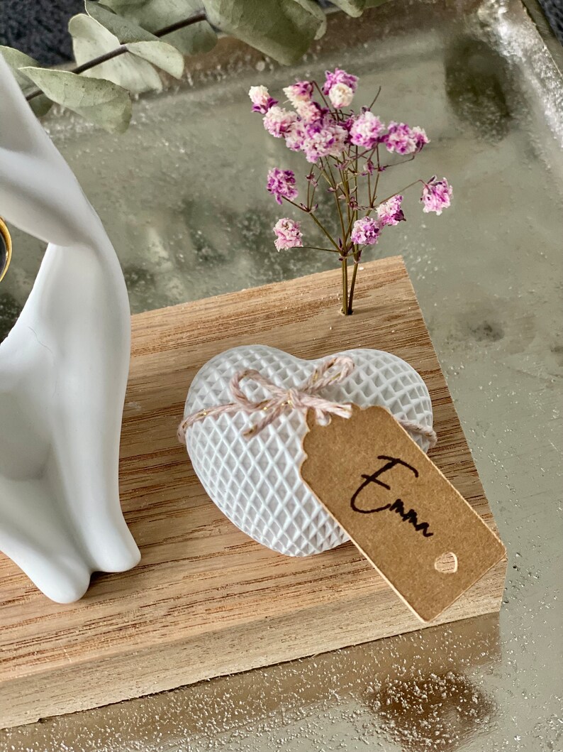 K&ouml;nnte beinhalten: Wei&szlig;e Keramikh&auml;nde halten ein kleines wei&szlig;es Herz mit einem braunen Etikett, auf dem "Emma" steht. Das Herz befindet sich auf einem Holzsockel mit einem kleinen Strau&szlig; rosa und wei&szlig;er Blumen.