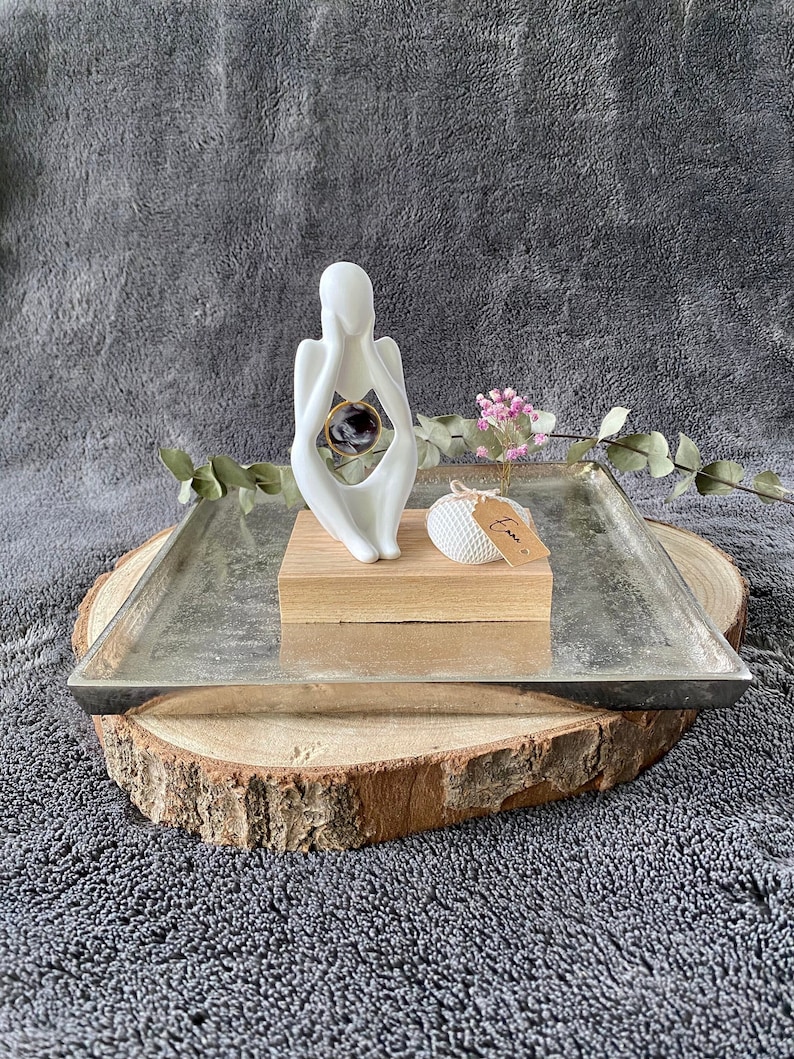 K&ouml;nnte beinhalten: Wei&szlig;e Keramikskulptur einer Person, die sich das Gesicht h&auml;lt, mit einer runden, durchsichtigen Glaskugel im Inneren der Skulptur. Die Skulptur steht auf einem Holzsockel und einem silbernen Tablett, das auf einer Holzscheibe liegt.
