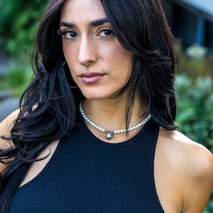 May include: A woman wearing a black ribbed tank top and a pearl necklace with a silver pendant. The necklace features a small, round, clear stone. The woman has long, dark hair and is looking at the camera.