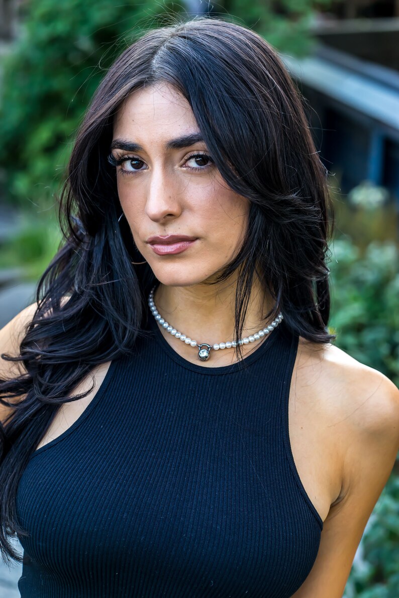 May include: A woman wearing a black ribbed tank top and a pearl necklace with a silver pendant. The necklace features a small, round, clear stone. The woman has long, dark hair and is looking at the camera.