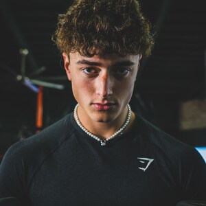 May include: A person wearing a black crew neck shirt with a white pearl necklace. The shirt has a white logo on the chest. The person has curly brown hair and is looking directly at the camera.