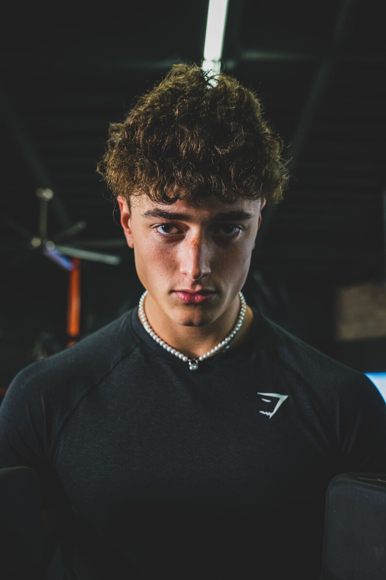 May include: A person wearing a black crew neck shirt with a white pearl necklace. The shirt has a white logo on the chest. The person has curly brown hair and is looking directly at the camera.