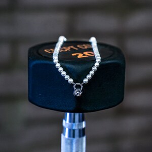 May include: A pearl necklace with a dumbbell pendant, featuring a clear gemstone. The necklace is displayed on a black dumbbell with the number 20 in orange. The background is a blurred brick wall.