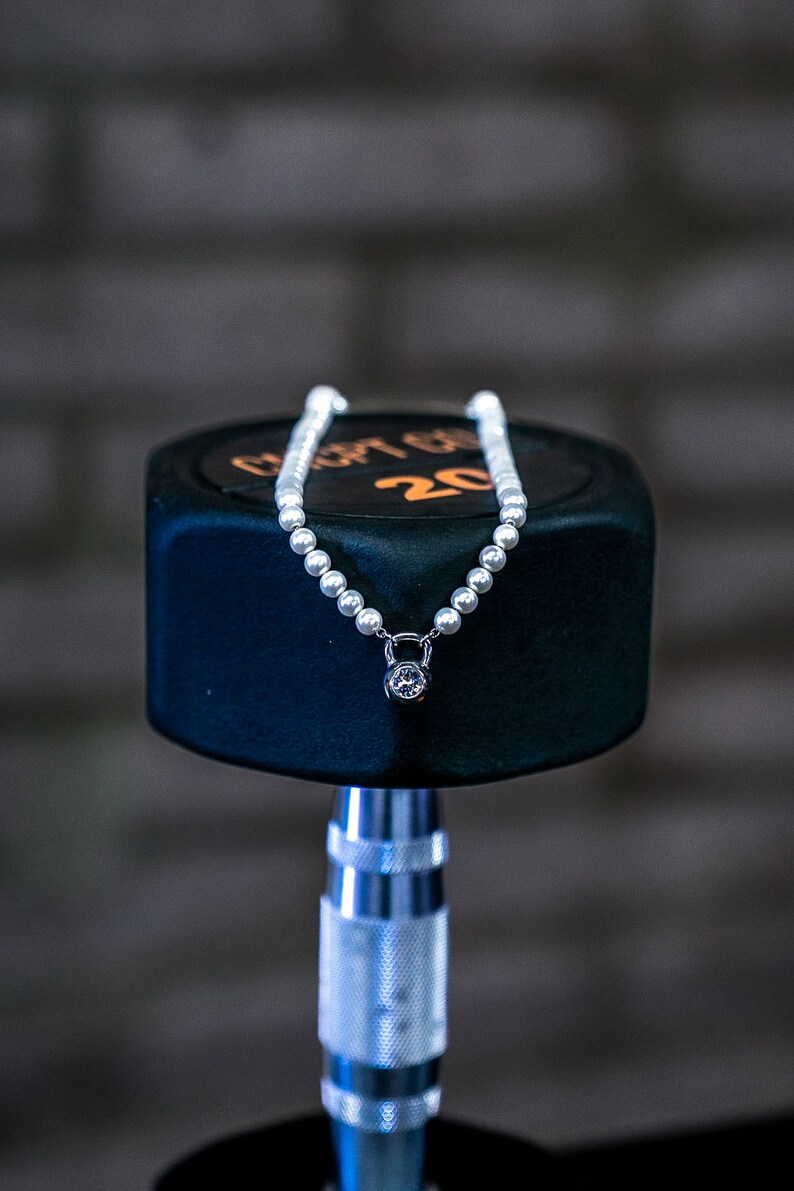 May include: A pearl necklace with a dumbbell pendant, featuring a clear gemstone. The necklace is displayed on a black dumbbell with the number 20 in orange. The background is a blurred brick wall.