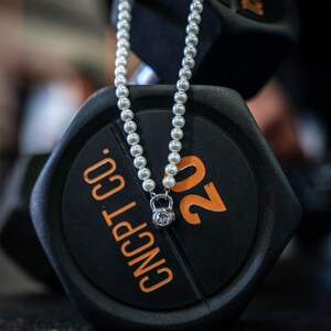 May include: A pearl necklace with a silver kettlebell pendant featuring a clear gemstone, resting on a black 20-pound weight. The weight has orange numbering and the text "CNCPT CO."