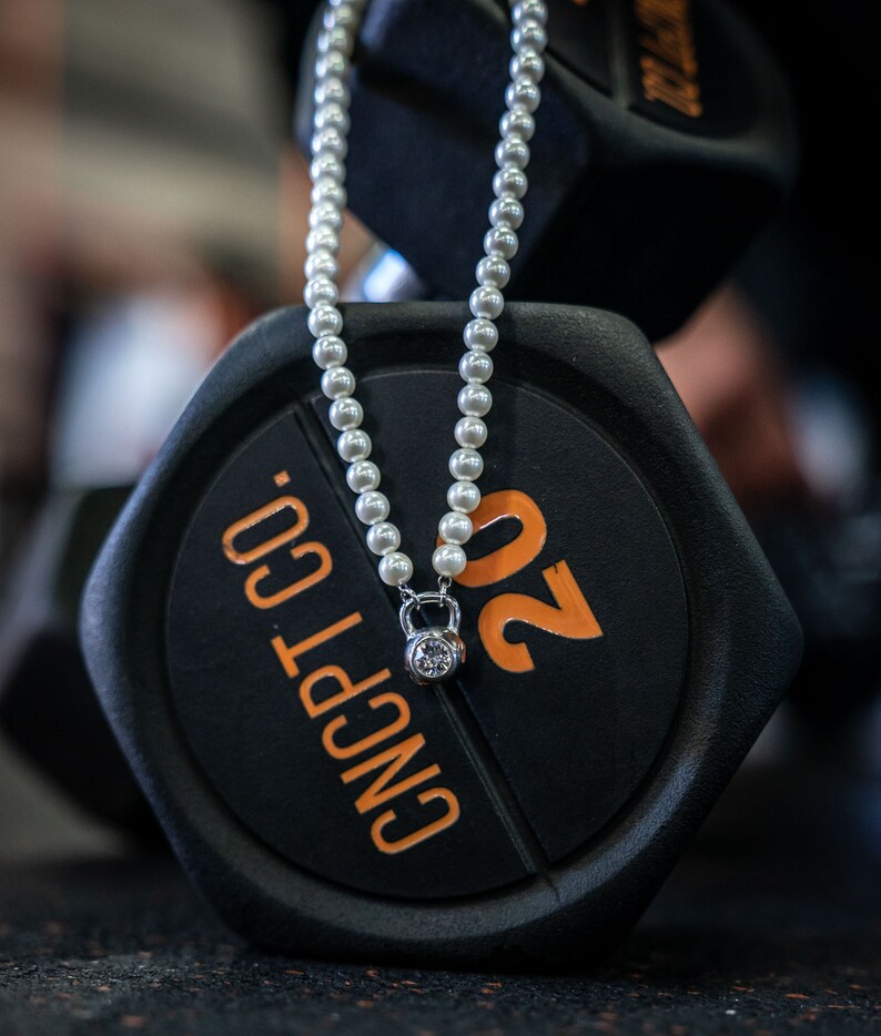 May include: A pearl necklace with a silver kettlebell pendant featuring a clear gemstone, resting on a black 20-pound weight. The weight has orange numbering and the text "CNCPT CO."