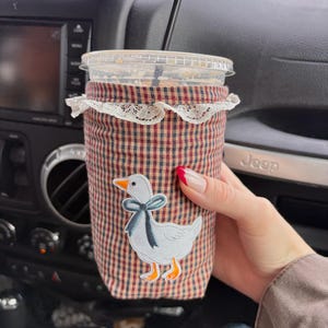 May include: A reusable coffee cup sleeve with a red and blue plaid pattern and a white lace trim. The sleeve features an embroidered goose with a blue bow. A clear plastic cup with a black straw is inside the sleeve.