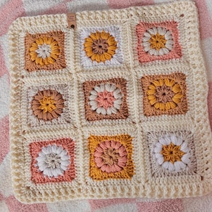 May include: A crocheted baby blanket with a cream background and colorful floral motifs in shades of pink, yellow, and brown. The blanket is made up of nine squares, each with a different floral design. The blanket has a label that reads "Little Joy Knits".