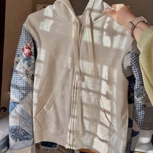 May include: Cream-colored zip-up hoodie with patchwork sleeves. The sleeves feature floral and blue gingham patterns. The hoodie has a drawstring hood and two front pockets. The garment is hanging on a white hanger.