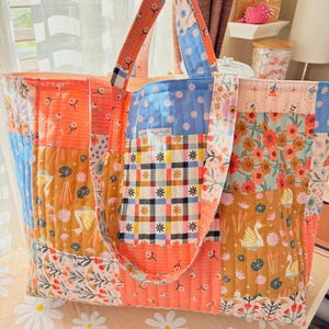 May include: A patchwork tote bag with a quilted design. The bag is made of various fabrics with floral and geometric patterns in shades of pink, blue, yellow, and brown. The bag has two long straps and a wide opening.