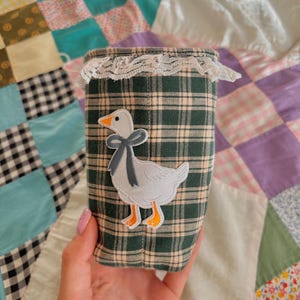 May include: A cylindrical fabric container featuring a dark green and beige plaid pattern. The top edge is trimmed with white lace. An embroidered patch of a white goose with an orange beak and feet, wearing a blue bow around its neck, is centered.