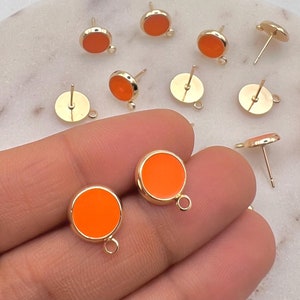 May include: A close-up of a hand holding a pair of orange and gold stud earrings. The earrings are round and have a smooth, glossy finish. The earrings are on a white surface.