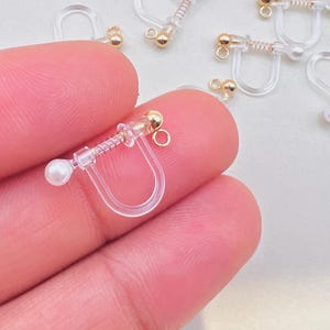 May include: Clear plastic clip-on earring converters with gold-colored accents and small faux pearls. The converters have a spring mechanism and a small loop for attaching earrings. Several converters are scattered on a light background.