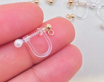 May include: Clear plastic clip-on earring converters with gold-coloured accents and small faux pearls. The converters have a spring mechanism and a small loop for attaching earrings. Several converters are scattered on a light background.