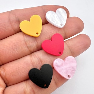 May include: Five heart-shaped acrylic charms in different colors. The charms are yellow, white, red, pink, and black. Each charm has a small hole at the top for attaching to a necklace, bracelet, or other jewelry.