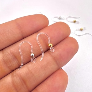 May include: Clear plastic earring hooks with silver and gold-colored beads. The hooks are curved and have a small loop at the bottom for attaching earrings. Several hooks are visible, some in focus, others blurred.