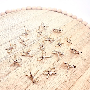 May include: A collection of gold-toned bow-shaped earring findings on a wooden surface. The findings are small and delicate, with a simple design.
