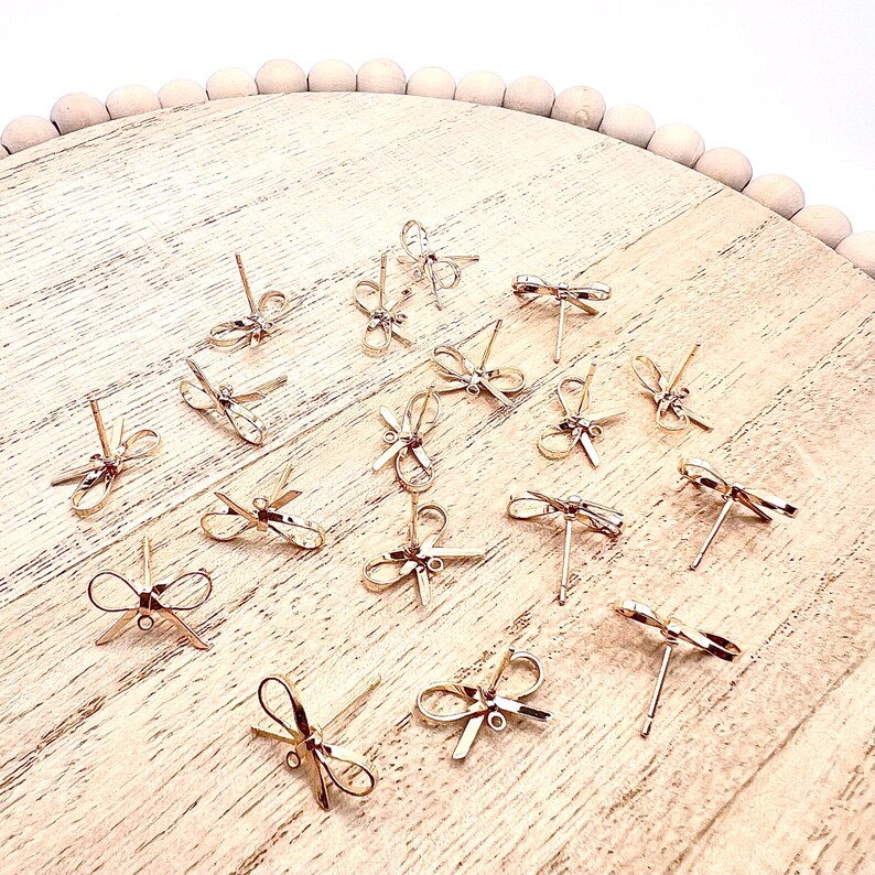 May include: A collection of gold-toned bow-shaped earring findings on a wooden surface. The findings are small and delicate, with a simple design.