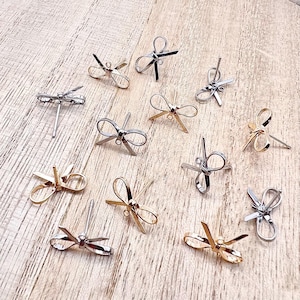May include: A collection of 12 silver and gold bow-shaped earring findings. The findings are arranged on a wooden surface.