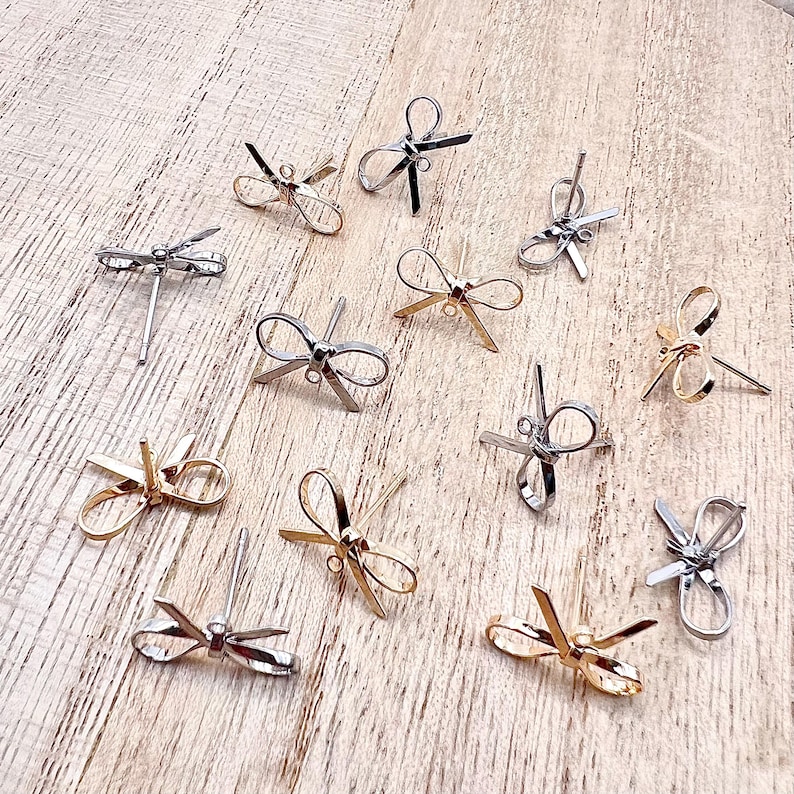 May include: A collection of 12 silver and gold bow-shaped earring findings. The findings are arranged on a wooden surface.