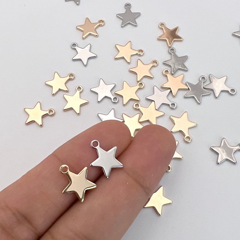 K&ouml;nnte beinhalten: Eine Sammlung von goldenen, silbernen und bronzenen sternf&ouml;rmigen Anh&auml;ngern mit Sprungringen. Die Anh&auml;nger sind auf einer wei&szlig;en Oberfl&auml;che verstreut.