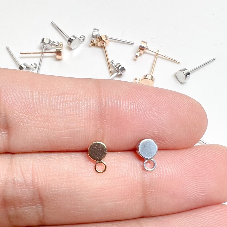 K&ouml;nnte beinhalten: Zwei Ohrring-Findings, einer golden und einer silbern, mit einer kleinen Schlaufe unten. Die Findings befinden sich auf einem Finger.