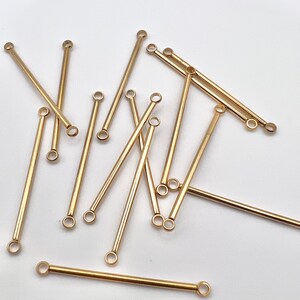 May include: A collection of gold-colored, straight bar connectors with small loops at each end. The connectors are scattered on a white surface, with varying lengths and orientations. These components are likely used in jewelry making or crafting.