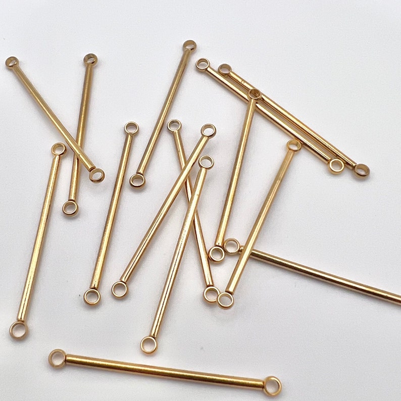 May include: A collection of gold-colored, straight bar connectors with small loops at each end. The connectors are scattered on a white surface, with varying lengths and orientations. These components are likely used in jewelry making or crafting.