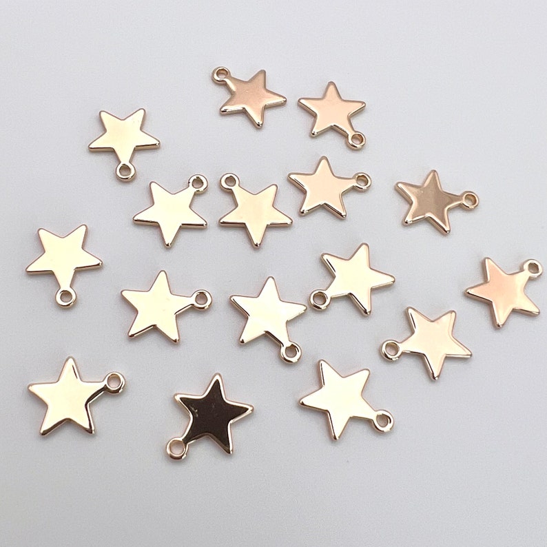 K&ouml;nnte beinhalten: Eine Sammlung von 16 goldfarbenen sternf&ouml;rmigen Charms mit einem Sprungring. Jeder Charm hat ein f&uuml;nfzackiges Sternendesign mit einer Schlaufe oben.