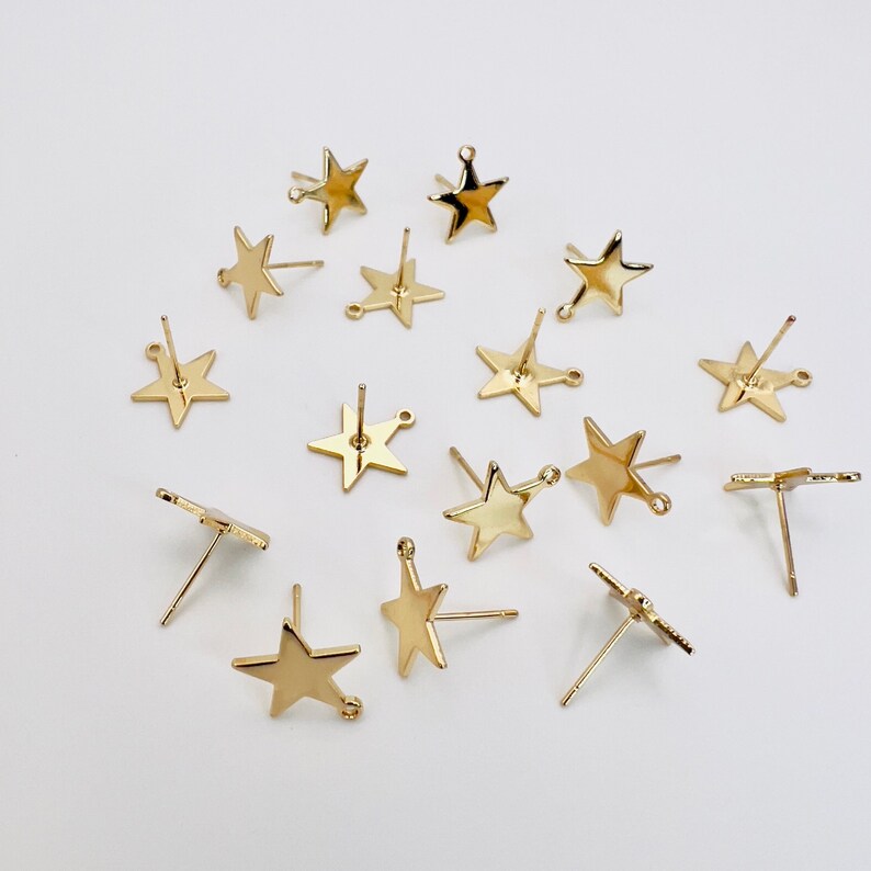 May include: A collection of gold-colored star-shaped earring posts. The earrings have a simple, minimalist design, with a post for insertion into the ear and a small loop for attaching charms. The earrings are scattered on a white surface.