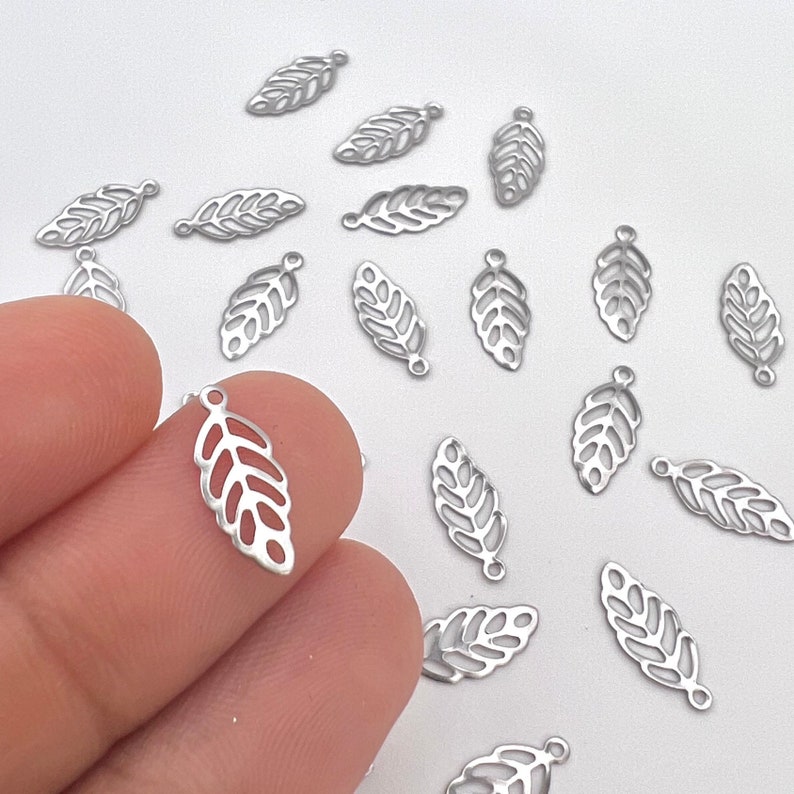 May include: A collection of silver metal leaf charms with a delicate cutout design. The charms are scattered on a white surface.