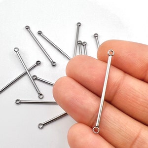 May include: A collection of silver-colored, straight bar jewelry findings with small loops at each end. Some are scattered on a white surface, while one is held in a person's hand. These findings are likely used for jewelry making, such as earrings or pendants.