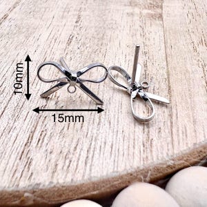 May include: Two silver bow-shaped earring findings with a jump ring and a post. The findings measure 15mm wide and 10mm tall.