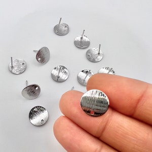 May include: Silver earring blanks with a textured surface and a small hole in the center. The earring blanks are round and have a post on the back.