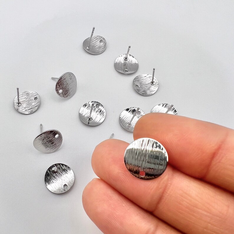 May include: Silver earring blanks with a textured surface and a small hole in the center. The earring blanks are round and have a post on the back.