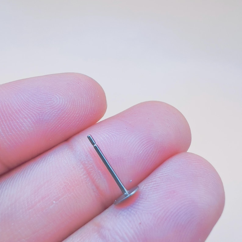 Pu&ograve; includere: Un singolo orecchino a bottone, piccolo, color argento, con un perno e un retro piatto e circolare. L'orecchino &egrave; tenuto tra due dita su uno sfondo neutro. Il perno dell'orecchino &egrave; dritto e sottile.