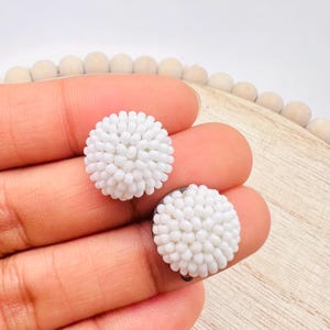 May include: Two white beaded stud earrings. Each earring is round and covered in small, white beads, creating a textured, floral-like appearance. The earrings are held in a person's hand, with a light wooden surface in the background.