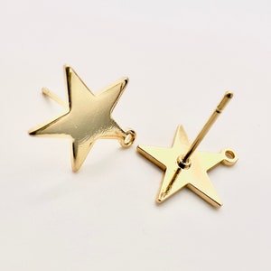 May include: Two gold-colored star-shaped stud earrings. Each earring has a post for insertion and a small loop at the bottom. The earrings are displayed on a white background.