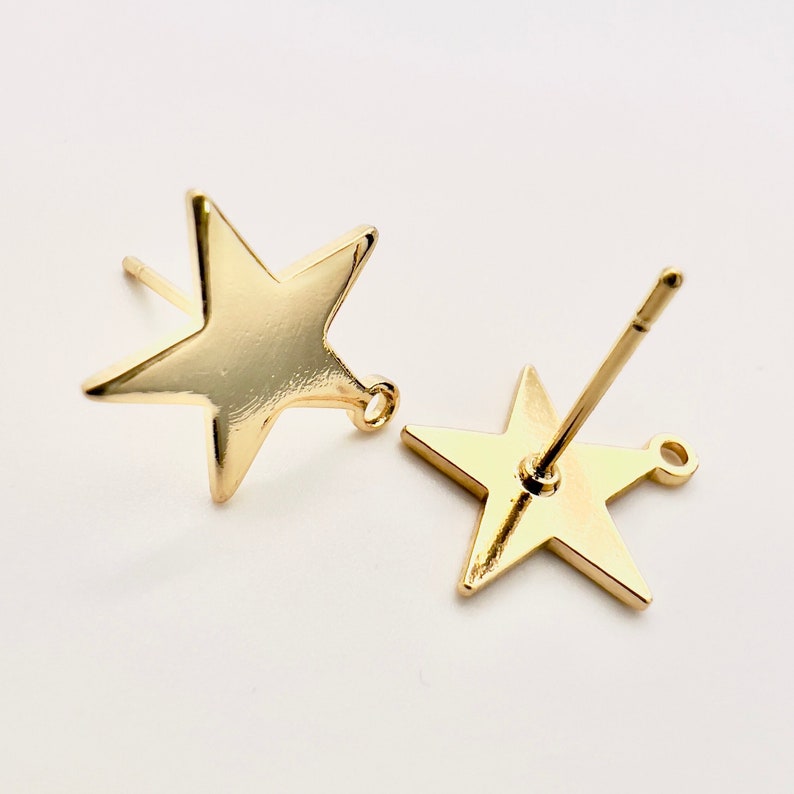 May include: Two gold-colored star-shaped stud earrings. Each earring has a post for insertion and a small loop at the bottom. The earrings are displayed on a white background.